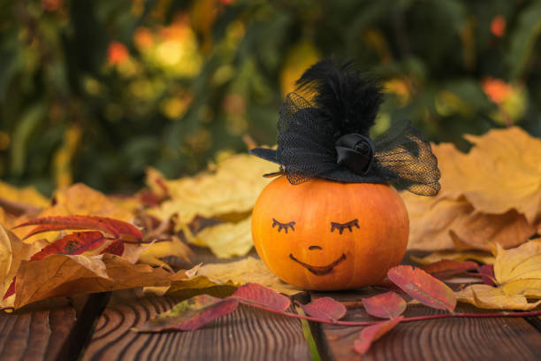 istock Pumpkin with a painted face in a black hat on a wooden table with leaves. 1278255229