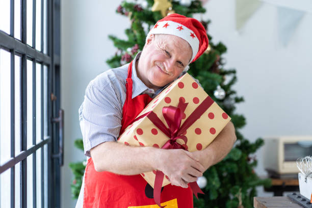 istock Smiling elderly man in red santa helper hat hugging gift box with Christmas tree blur background at home. Merry Christmas, X-mas, Winter, Happy people concept 1278259389