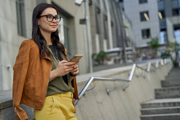 vista lateral de uma jovem mulher branca bonita e elegante usando seu smartphone e sorrindo enquanto passava um tempo ao ar livre, parada na rua da cidade - shatting - fotografias e filmes do acervo