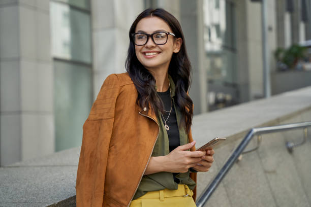 ritratto di una giovane donna bella e alla moda che indossa occhiali con in mano lo smartphone, guarda da parte e sorride mentre si trova sulla strada della città - shatting foto e immagini stock