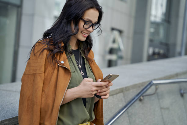 widok z boku młodej pięknej i modnej kobiety noszącej okulary za pomocą smartfona, rozmawiając z przyjacielem i uśmiechając się stojąc na niewyraźnym tle miejskim - shatting zdjęcia i obrazy z banku zdjęć