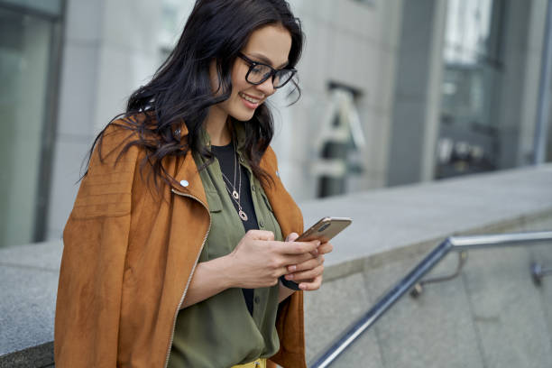 vista lateral de uma jovem linda e elegante mulher usando óculos usando seu smartphone, conversando com a amiga e sorrindo enquanto estava contra o fundo urbano desfocado - shatting - fotografias e filmes do acervo