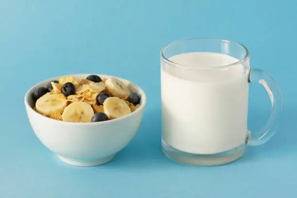 Oat granulated crispy flakes in bowl with slices of banana, blueberries and with  full  cup of fresh milk on a blue background, close-up. Concept - healthy food, breakfast