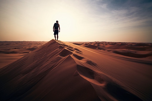 Young female sandboarding in The Sahara Desert during the sunset. She is standing on a sand dune, holding sandboard and watching the sunset over desert.