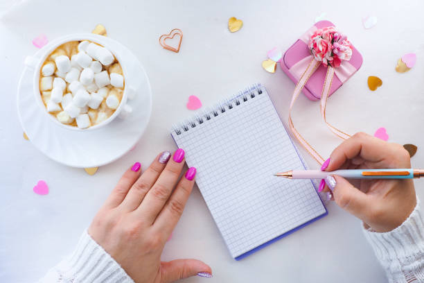 istock female hands with a beautiful manicure write with a pen in a notebook and a white cup with coffee with marshmallows, a pink gift box and heart-shaped sweets. flat layout, top view, lifestyle, 1278272352