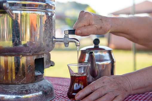 Samovar and Turkish tea enjoyment at picnic. Woman's hand pouring tea from samovar into glass. Traditional drinks and holiday concepts. Horizontal close-up with copy space.