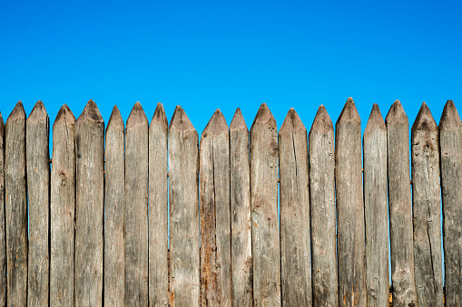 Fence made of sharp wooden stakes against the blue sky. Wooden fence vertical logs pointed against the sky protection against invaders and wild animals