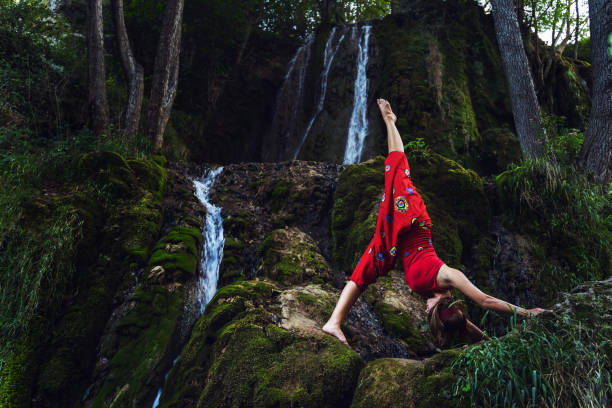 ritratto frontale di donna caucasica adulta di 30 anni seduta su roccia nella natura di fronte alla cascata in abito rosso che si estende in posa yoga asana - spiritualità interiore pace e relax concetto - waterfall zen like women meditating foto e immagini stock