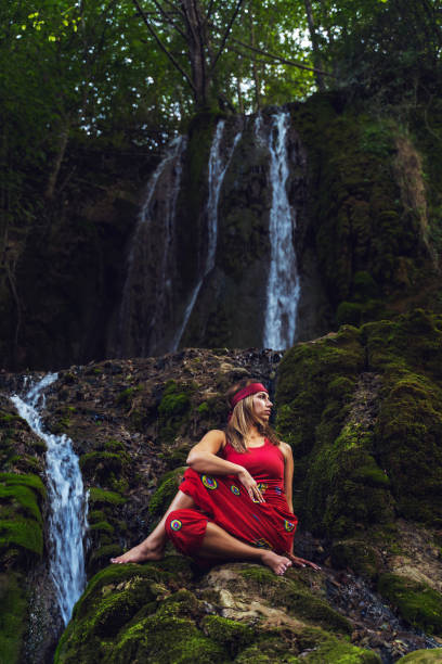 ritratto frontale di donna caucasica adulta di 30 anni seduta su roccia nella natura di fronte alla cascata in abito rosso che si estende in posa yoga asana - spiritualità interiore pace e relax concetto - waterfall zen like women meditating foto e immagini stock