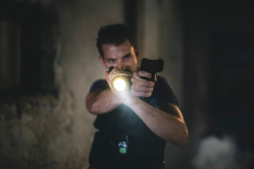 Close-up of young Caucasian police officer in early 30s aiming flashlight and pistol into darkness of run-down urban location.