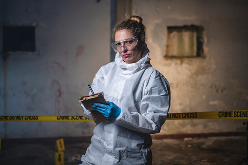 Female crime scene investigator in late 30s standing outside evidence area and looking at camera while making notes.