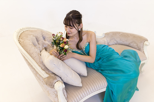 Portrait of young woman in turquoise blue dress sitting on couch and looking at bouquet