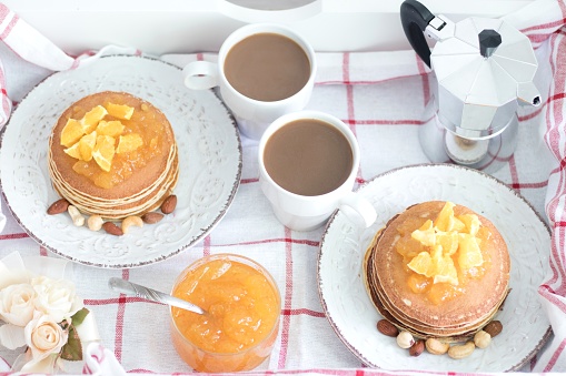 Romantic traditional breakfast for two on a tray. American pancakes with orange jam and nuts on vintage plates and 2 white coffee cups with Italian metal coffee pot on a checkered cloth. Top view