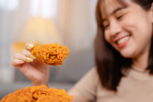 Hungry young woman eating junk food Fried Chicken and French Fries for lunch by ordering delivery, relaxing at home on holiday. unhealthy meal, obesity risk.