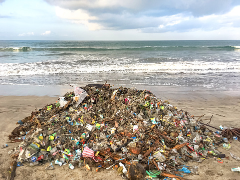 Close up shot of plastic garbage pile up in Bali beach, on the shoreline with a background of the ocean waves and blue cloudy sky