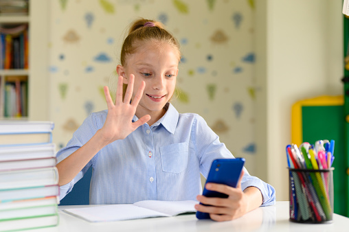 A girl holds a smartphone and communicates on the Internet via video communication. Greets his interlocutor with a gesture of his hand and smiles.