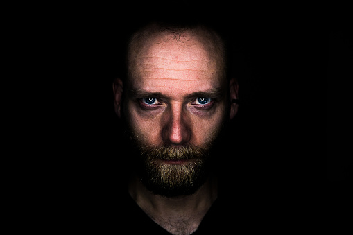 A man looks seriously into the camera from the darkness. His look is threatening and determined.