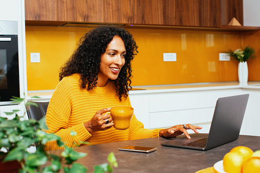 Retrato de una mujer mediana con el pelo rizado negro sonriendo mientras usa la computadora portátil en la cocina photo