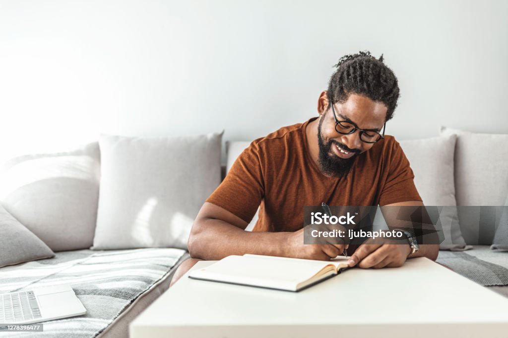 Control your budget before it controls you Image of happy African man with notepad and pen sitting on sofa. Writing - Activity Stock Photo