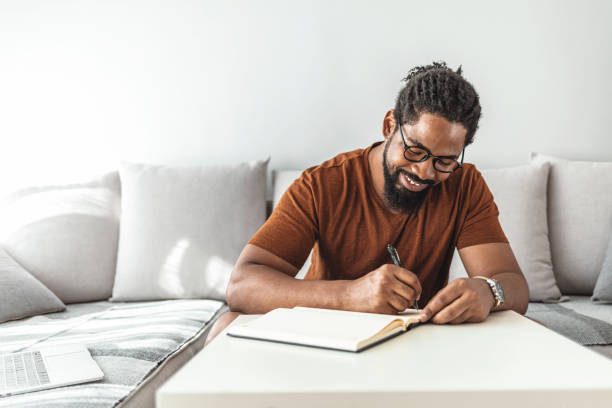 Control your budget before it controls you Image of happy African man with notepad and pen sitting on sofa. writer stock pictures, royalty-free photos & images