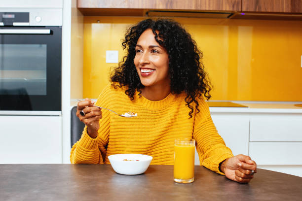 verticale d’une femme moyenne avec le sourire bouclé noir tout en mangeant des céréales de petit déjeuner dans la cuisine - yellow and black photos et images de collection