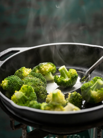 Fresh broccoli spears being sauteed with the use of a cooking utensil, stirring the vegetable during cooking, in a heavy iron pan on a gas stove against a turquoise colored background. Shot from a high angle above, good copy space at the top of the image. very shallow depth of field with the shallow focus being on the spears in the center of the pan.