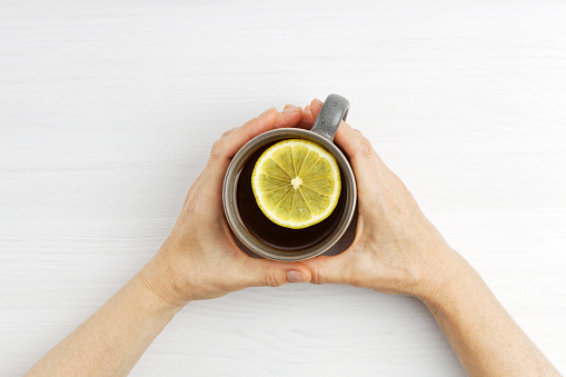 Female hands hold a warm cup of tea with a slice of lemon . Top view