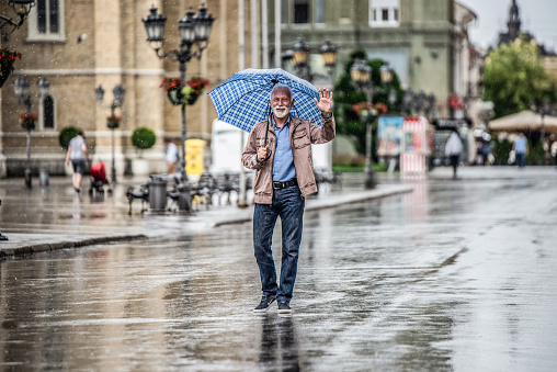 Happy senior man walking on rainy day under umbrella in the city. Looking at camera.