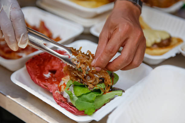 Close up shot of male hands with protective gloves putting caramelized onion above the bun as he preparing vegan burger ,on top of a takeout box