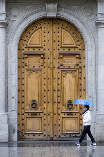 a woman walks with an umbrella in profile with a protective mask in front of a porte-cochere