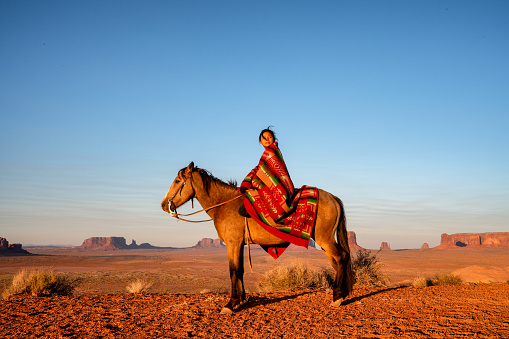 Niña Navajo de doce años sentada en lo alto de su caballo de color Bay Brown frente a los famosos Buttes en el Parque Tribal Monument Valley en el norte de Arizona EE.UU. al atardecer con una manta india photo