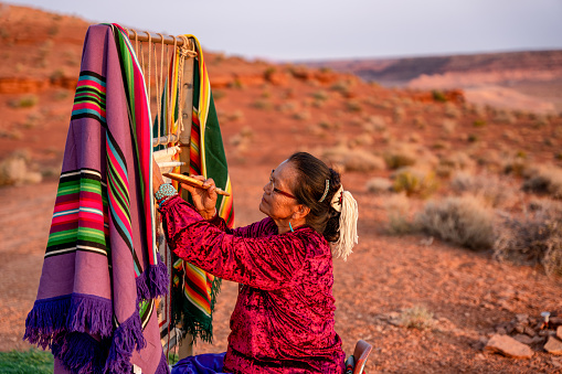 Anciana mujer navajo tejiendo una manta tradicional o alfombra en un telar nativo americano auténtico en el desierto al atardecer cerca del parque tribal Monument Valley en el norte de Arizona photo