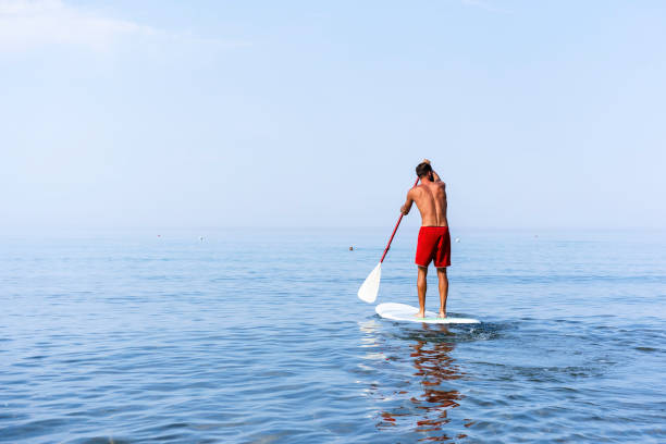 istock Athletic young man paddling on a sup board on the quiet sea - Stand up paddle boarder training on a rowing board on a flat calm sea - Back view and copy space for text 1278438403