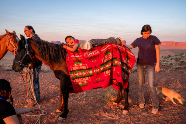 carino tre anni vecchio nativo americano indiano navajo ragazzo sdraiato sul retro di un cavallo come la sua famiglia si prepara a cavalcare in arizona settentrionale al monument valley tribal park sulla riserva navajo a crepuscolo - cherokee foto e immagini stock