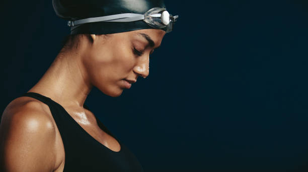 Woman swimmer looking tired Close-up of a woman swimmer looking tired after a race. Female wearing black swimsuit, swimming cap and goggles on dark background. black women in bathing suits stock pictures, royalty-free photos & images