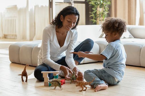Smiling beautiful young african ethnicity woman sitting on warm wooden floor with adorable joyful small kid boy, playing toys in modern living room, children daycare domestic activity indoors.