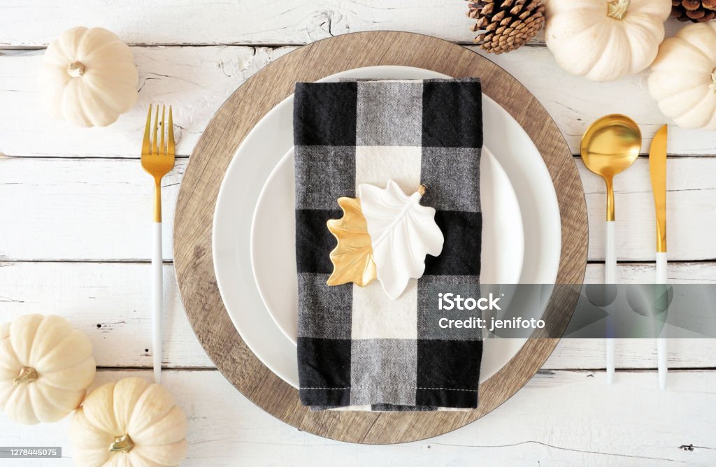 Autumn harvest or thanksgiving dinner table setting. Top view on a white wood background. Autumn harvest or thanksgiving dinner table setting with plates, flatware, buffalo plaid napkin, pumpkins and decor. Top view on a white wood background. Place Setting Stock Photo
