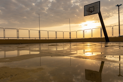 Empty and wet basketball court after the rain with puddles reflecting sunlight in the background.