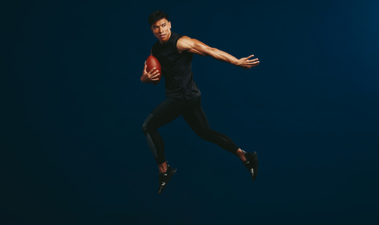 American football player holding ball and jumping against dark background. Sportsman practising football.