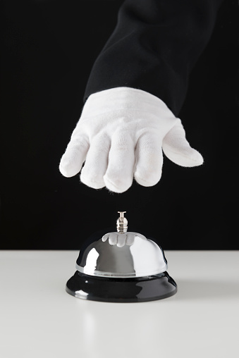 Hand with white glove is ringing a service bell in front of black background. The service bell is on a white table.