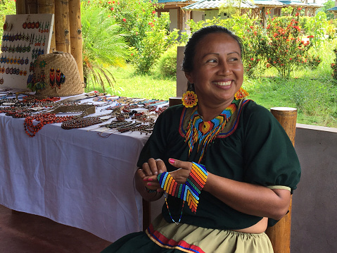 Nueva Loja, Sucumbios / Ecuador - September 2 2020: Indigenous woman of Cofan nationality with green dress smiling while weaving handicrafts sitting on a chair at her home in the Amazon rainforest