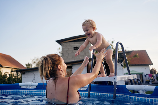 Beautiful innocent cheerful baby boy having fun at the pool with his young mother.