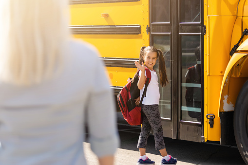 A yellow school bus is parked with its flashing Stop sign folded out as a small group of students cross in front.  The students are dressed casually and have backpacks on as they make their way to board the bus.