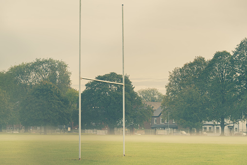 Rugby goal posts on a playing field on a misty evening.  Across the playing field is a row of trees, fence and a residential street.  Belfast, Northern Ireland.