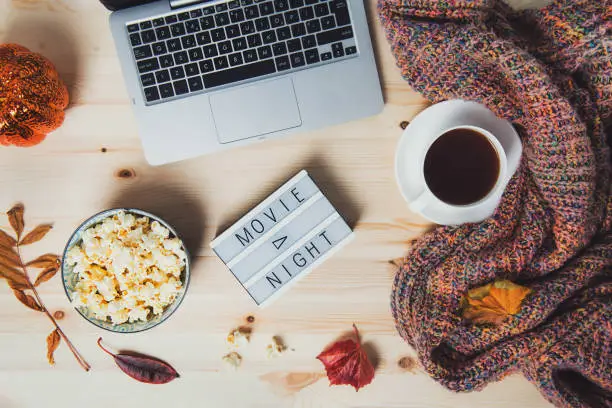 Top view Movie night concept. Flat lay composition with Movie night message on the board, laptop, popcorn bowl, decorative pumpkin, fallen leaves, a cup of tea, and warm plaid on wooden background