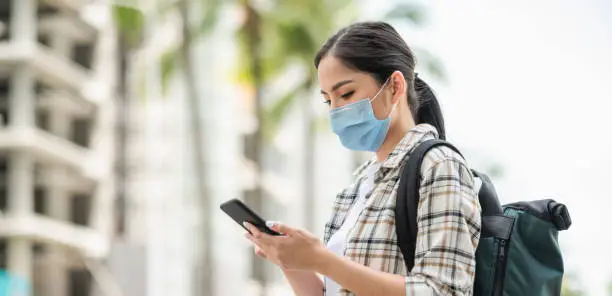 Photo of Young Asian beautiful woman travelers use her mobile phones to find travel locations and call for taxi transportation. She wearing protection mask for new normal safety travel trips.