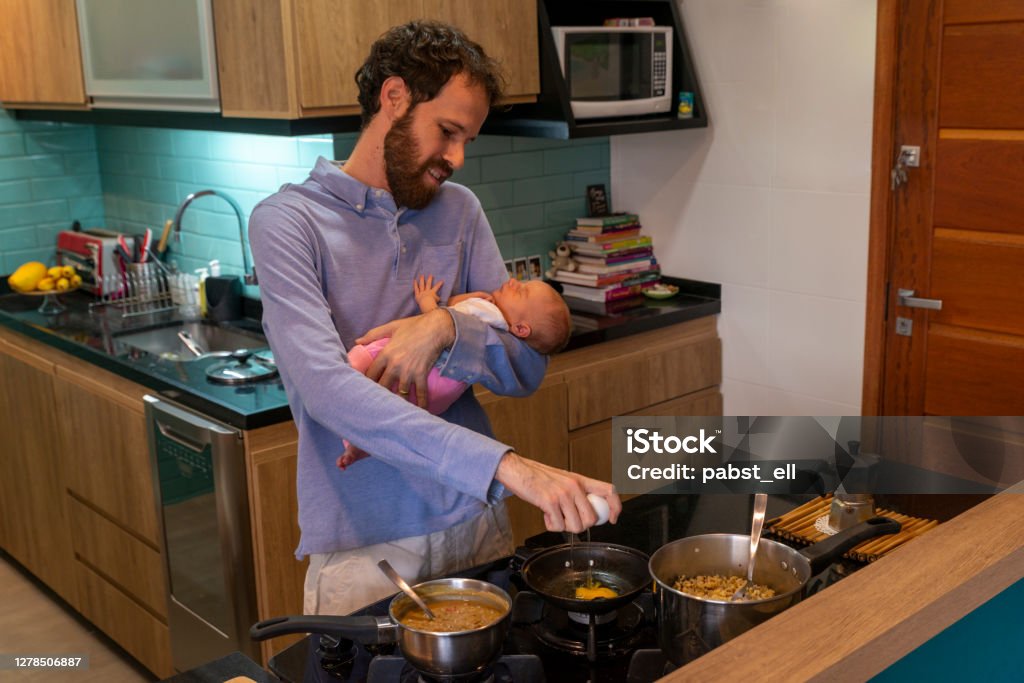Father holding baby while cooking vegetarian Father on the kitchen holding his baby daughter while cooking vegetarian food. Cooking Stock Photo