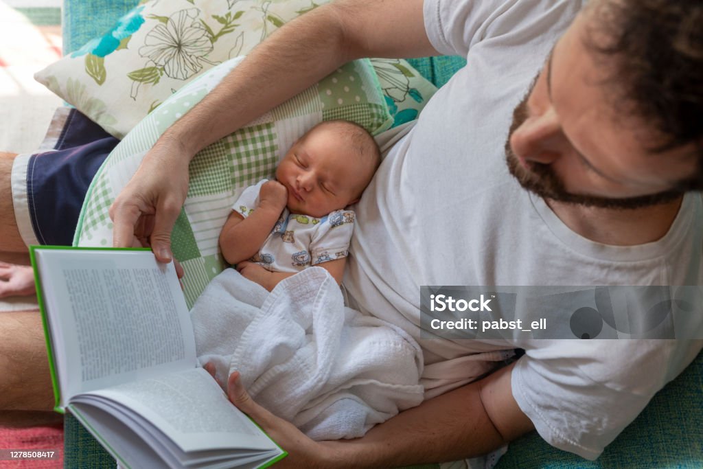Father reading paper book with baby on lap Father reading a book on the sofa with his baby daughter sleeping on his lap on the sofa. Paternity Leave Stock Photo