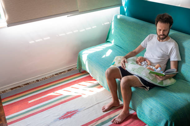 Father on paternity leave with baby reading at home Father reading a book on the sofa with his baby daughter sleeping on his lap on the sofa. paternity stock pictures, royalty-free photos & images