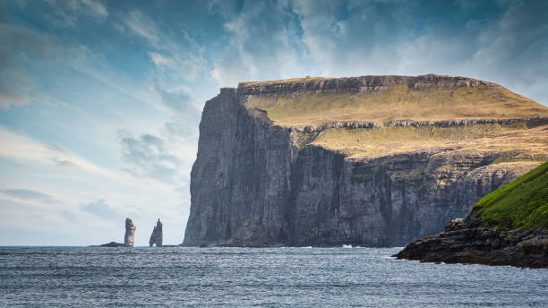 Risin og Kellingin Sea Stacks Faroe Islands Eysturoy Panorama Risin og Kellingin Sea Stacks Panorama. Risin and Kellingin Seas Stack - in the North Atlantic Ocean under blue summer skyscape. Risin og Kellingin means 'The Giant and the Witch' and references to an old legend about the sea stack origins. Risin is the 71m high stack further from the coast (left), and the Kellingin is the 68m stack nearer land (middle). These numbers gives a relation how the dimensions how tall and gigantic the fjord cliff coast is (on the right). Northern Coast of the Island of Eysturoy close to the town of Eiði. Eysturoy Island, Faroe Islands, Kingdom of Denmark, Nordic Countries, Europe eysturoy photos stock pictures, royalty-free photos & images